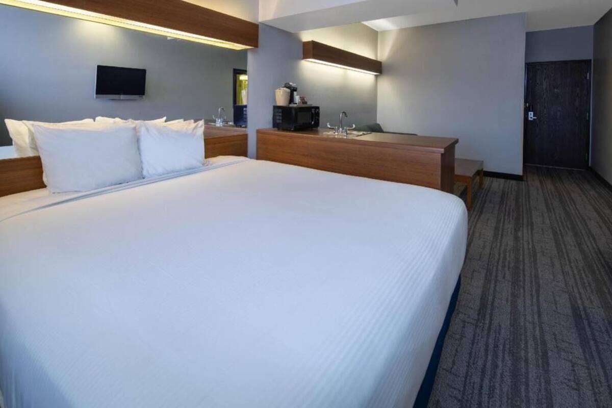 A hotel room with a large bed, a wooden nightstand, a built-in desk with a sink, and a television mounted on the wall.