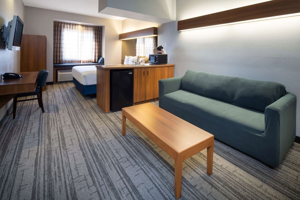 A hotel room with a bed, a desk with a chair, a sofa, a small kitchenette, and a TV mounted on the wall.