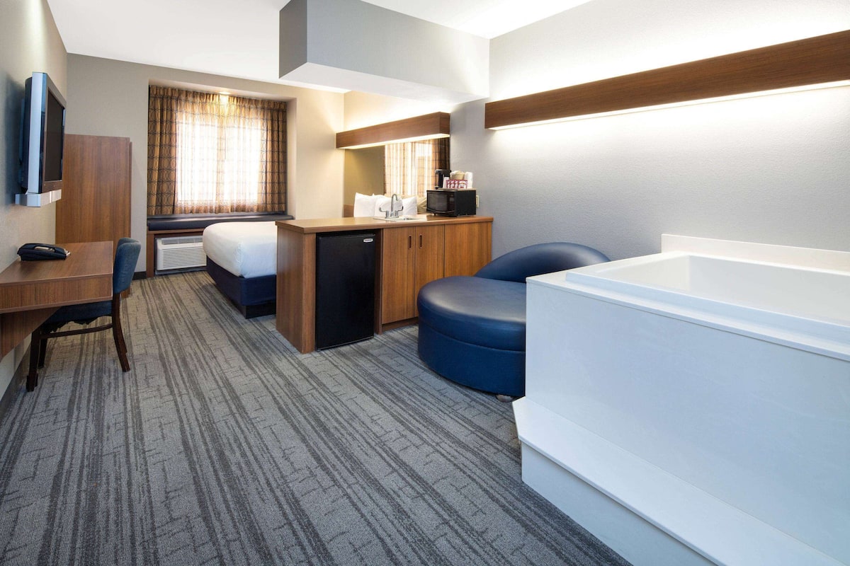A hotel room with a bed, desk, chair, microwave, and a large bathtub.