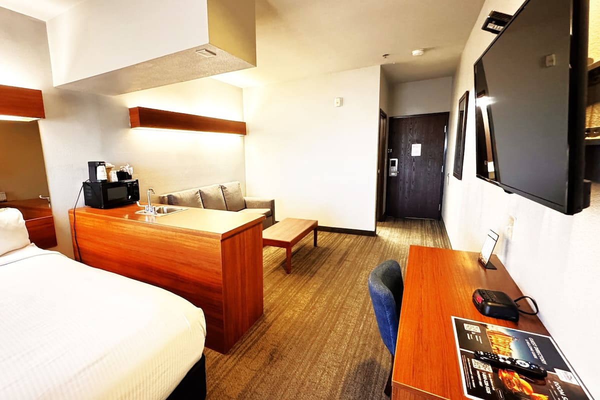 A hotel room with a bed, a wooden desk, a small kitchenette, a sofa, and a TV mounted on the wall.