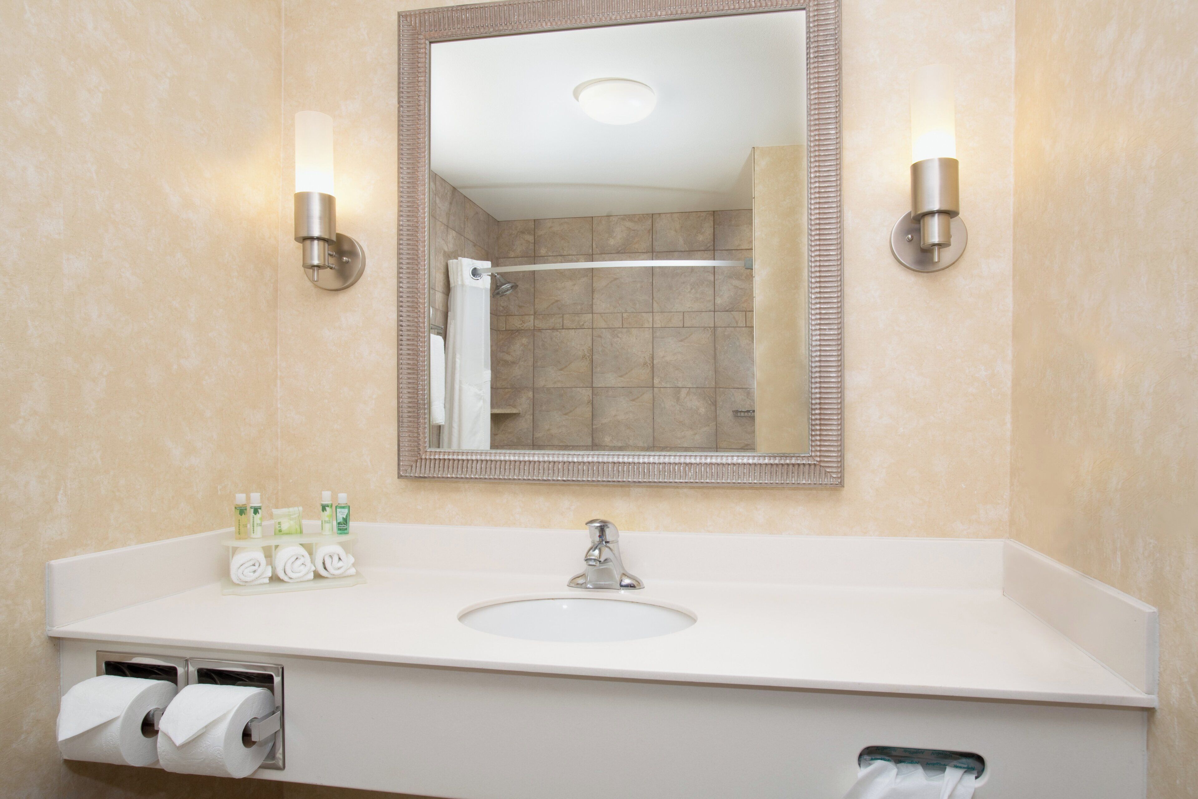 Baño con lavamanos, espejo, dispensador de toallas y una ducha visible en el reflejo.