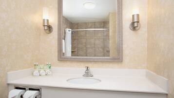 Baño con lavamanos, espejo, dispensador de toallas y una ducha visible en el reflejo.