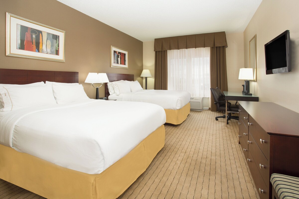 A hotel room with two beds, a desk, a chair, a television, and artwork on the walls.