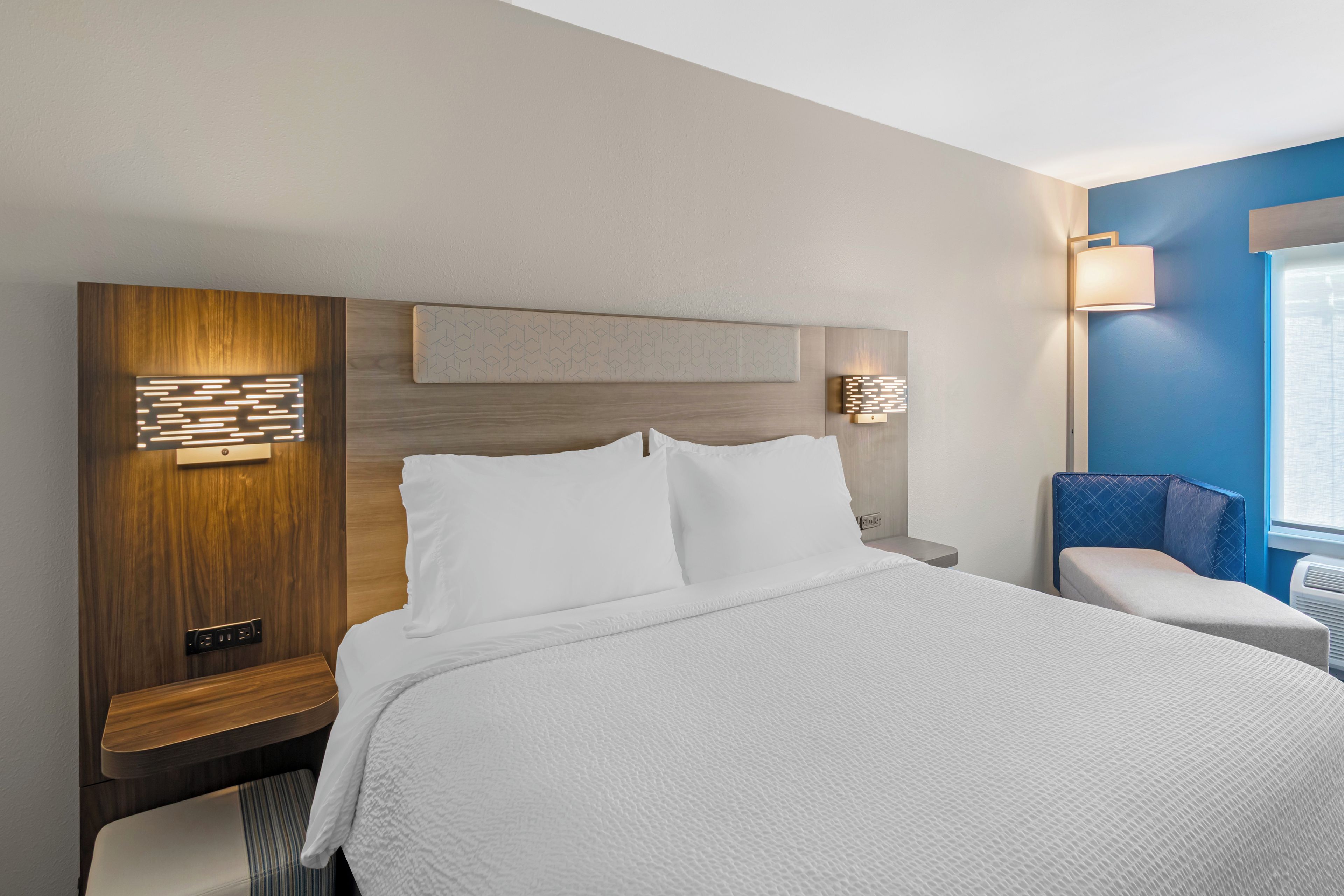 A hotel room with a large bed, a wooden headboard, two bedside tables, a wall-mounted light fixture, and a window with blinds.