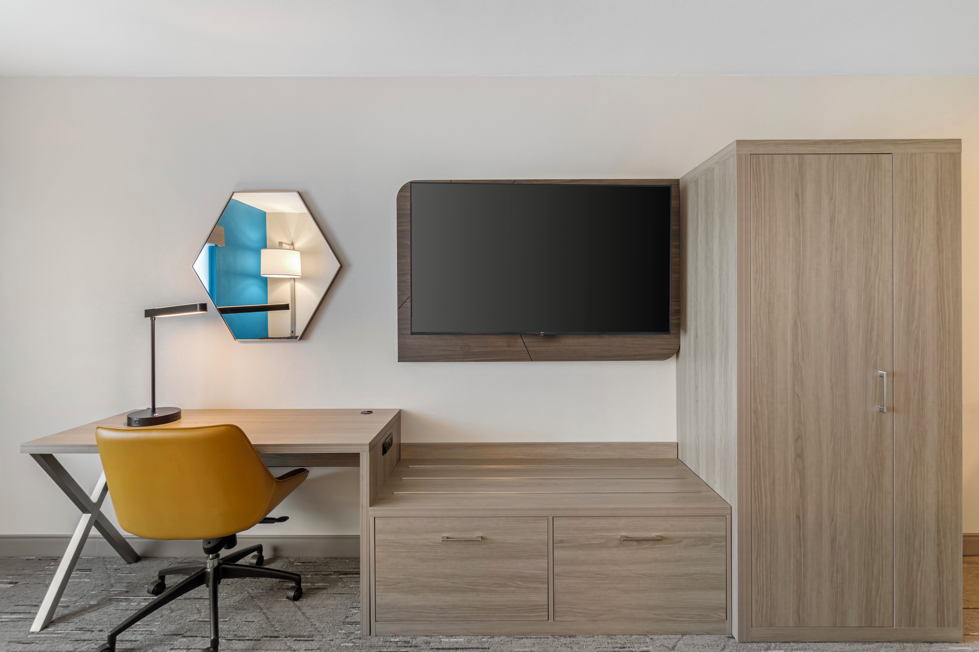A modern hotel room with a wooden desk, a yellow chair, a wall-mounted TV, and a wardrobe.