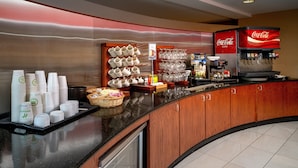 A hotel breakfast area with a Coca-Cola dispenser, stacks of white cups, and a variety of glassware.