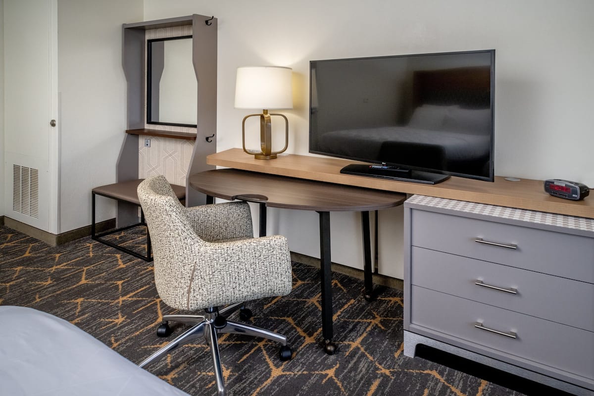 A hotel room with a bed, a desk with a television, a chair, a dresser, and a mirror.