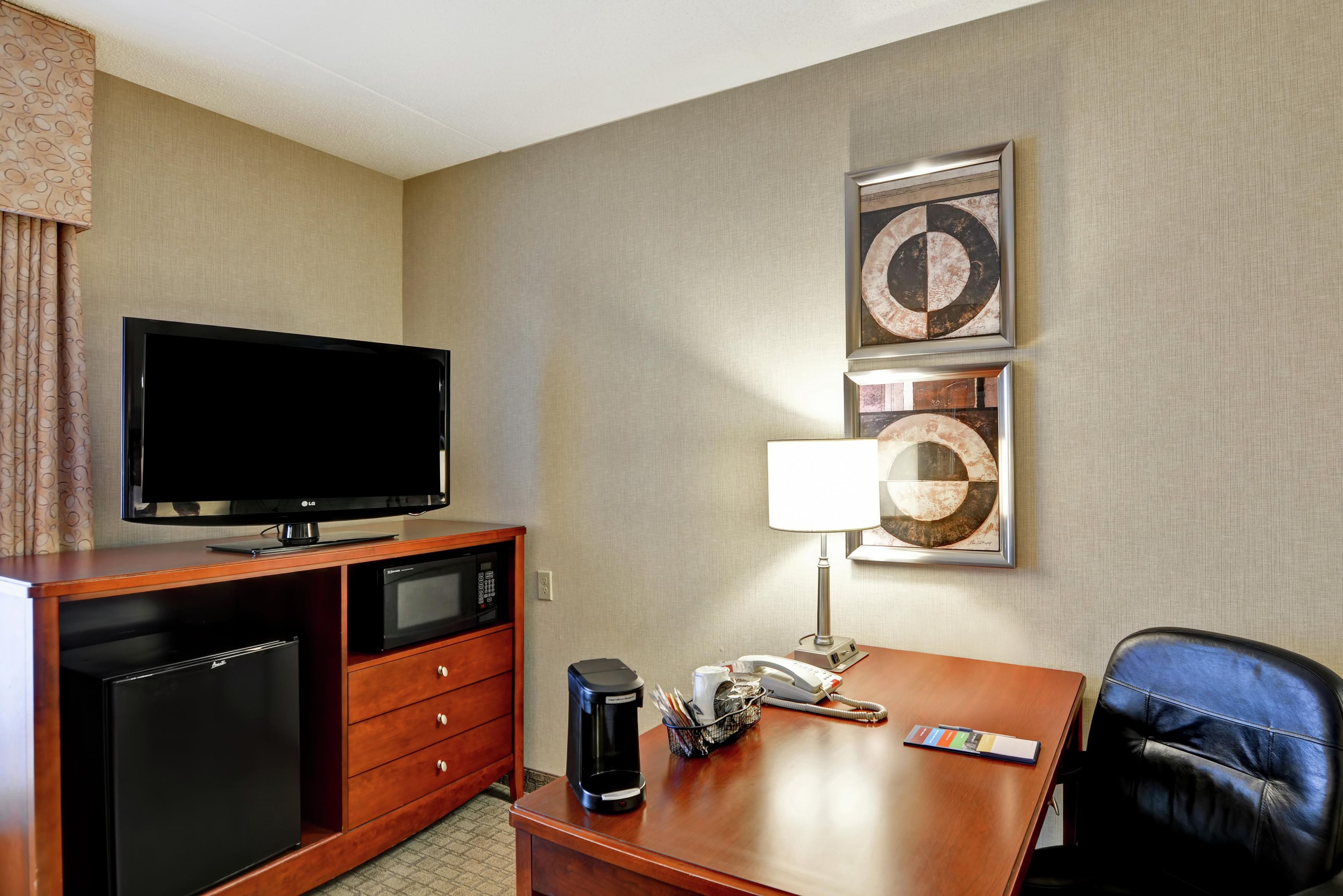 Quarto de hotel com televisão de tela plana, cafeteira, telefone, luminária e uma obra de arte emoldurada na parede.