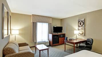 Quarto de hotel com cama, escrivaninha, cadeira, televisão, minibar e janela com cortinas.