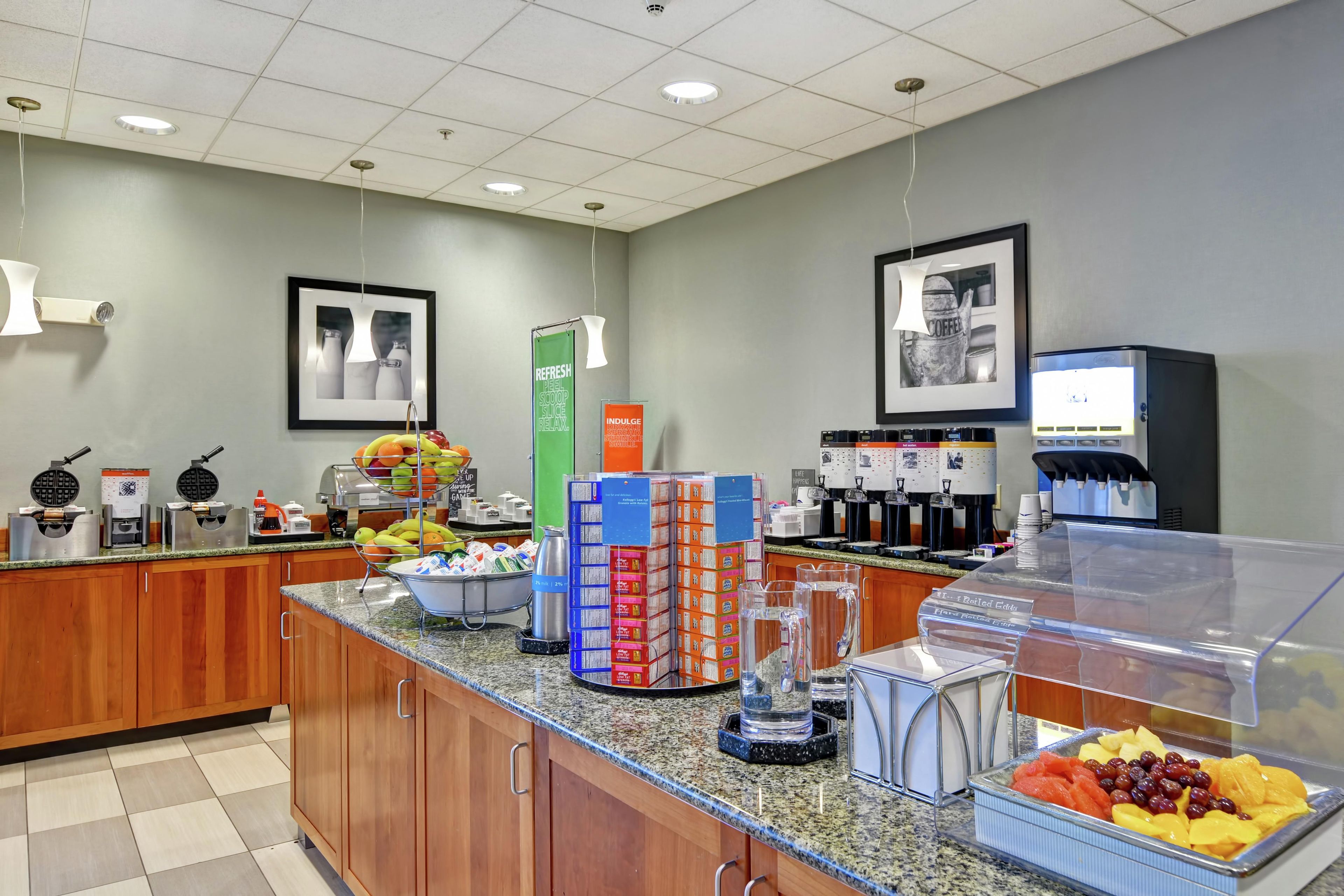 A self-service breakfast area with waffle makers, fruit, coffee machines, and a water dispenser.