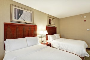 A hotel room with two beds, a nightstand with a lamp, and two framed artworks on the wall.