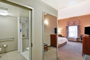 A hotel room with a shower, toilet, bed, bedside table, chair, and a TV.