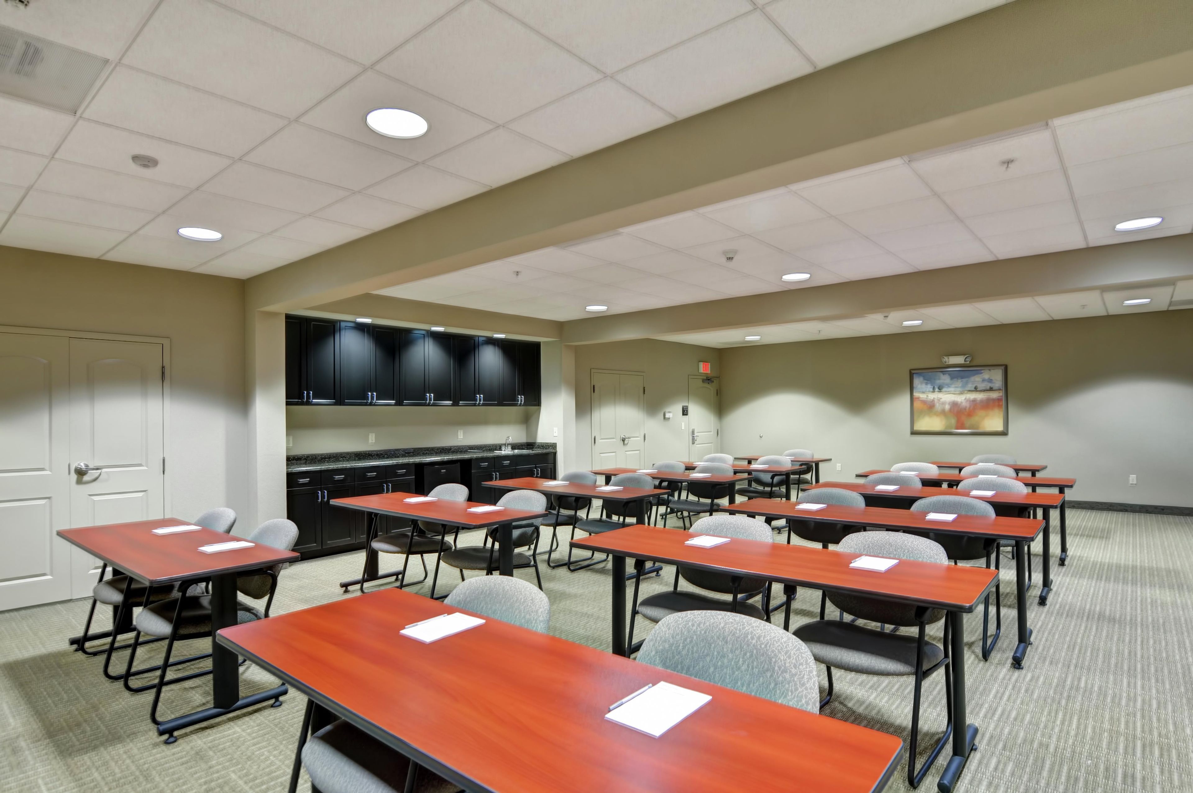 A conference room with multiple tables and chairs, a kitchenette area, and a painting on the wall.