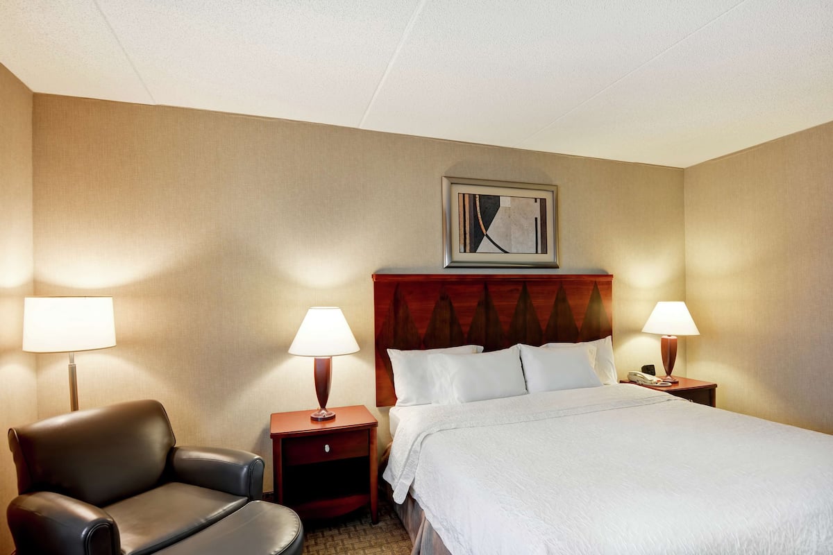 Habitación de hotel con cama, dos mesitas de noche con lámparas, una silla y cabecera con un cuadro enmarcado.