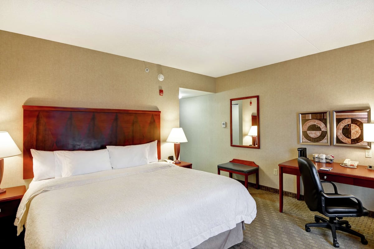 Habitación de hotel con una cama grande, un escritorio, una silla, una lámpara, un espejo y cuadros en la pared.