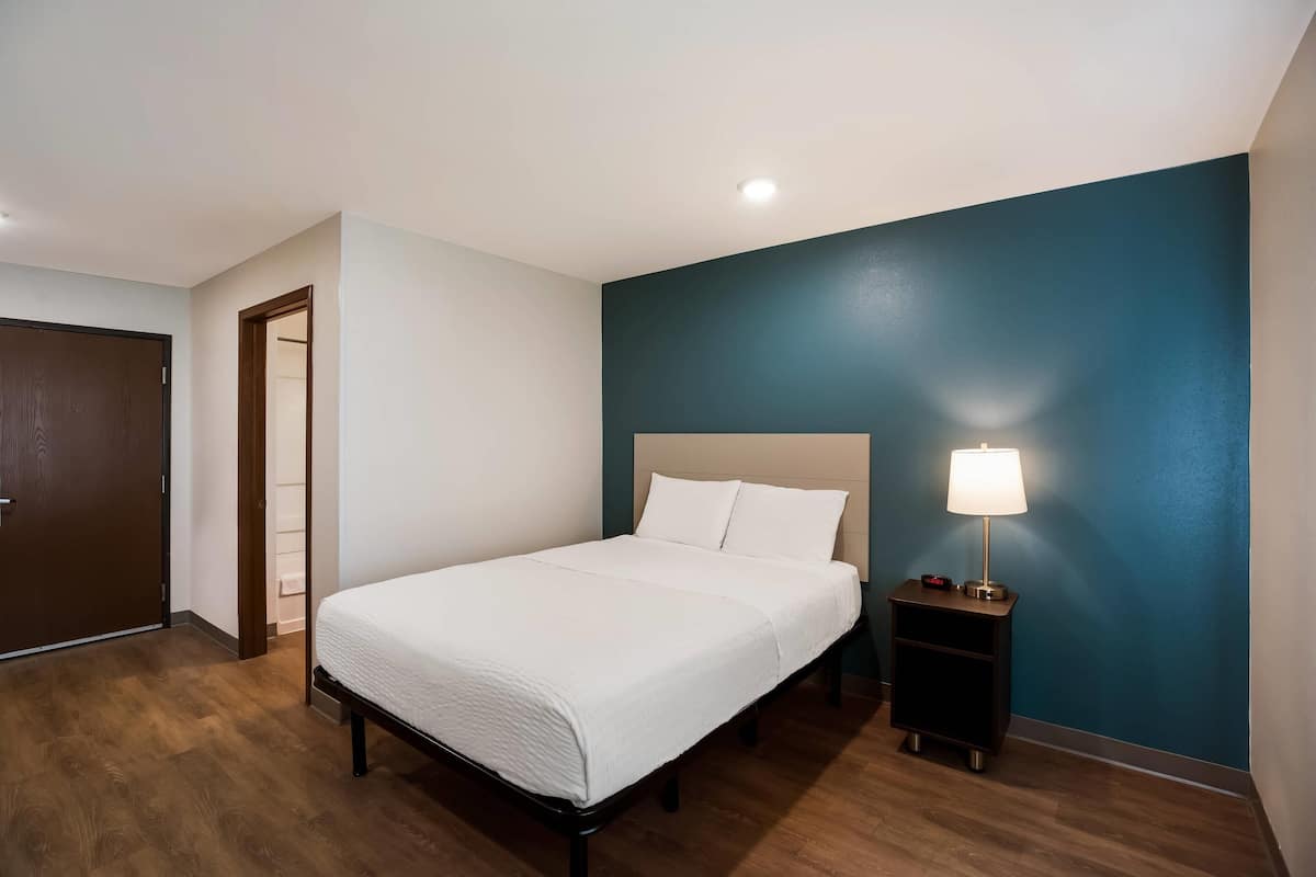 Un lit bien fait dans une pièce dont un mur est peint en bleu, avec un plancher en bois, une table de chevet avec une lampe et une porte.