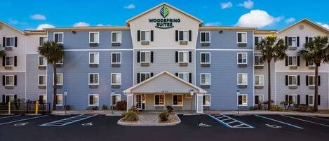 A Woodspring Suites hotel with a central entrance, multiple floors, and a parking lot with handicapped parking spaces.
