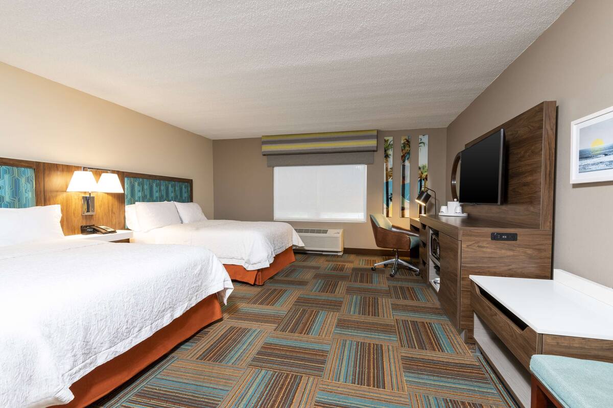 A hotel room with two beds, a desk, a TV, and a window.