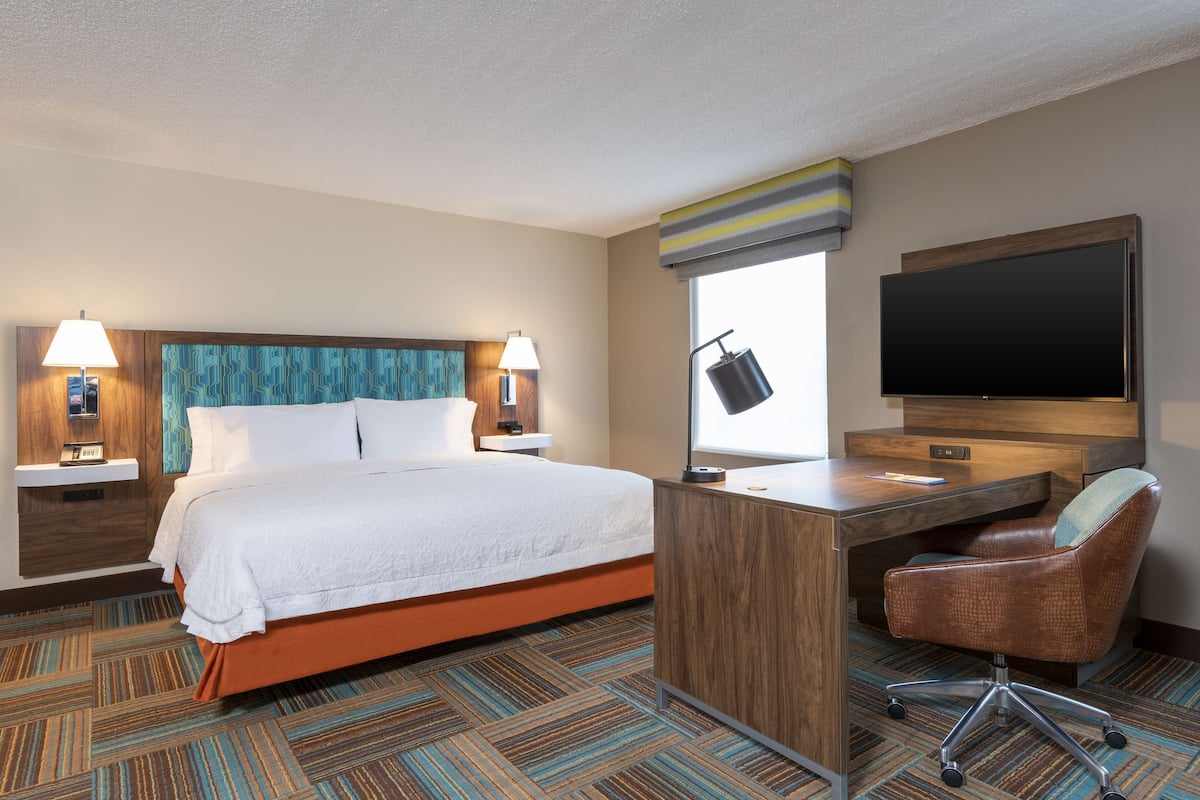 A hotel room with a large bed, a desk, a chair, a TV, and a window with a view.