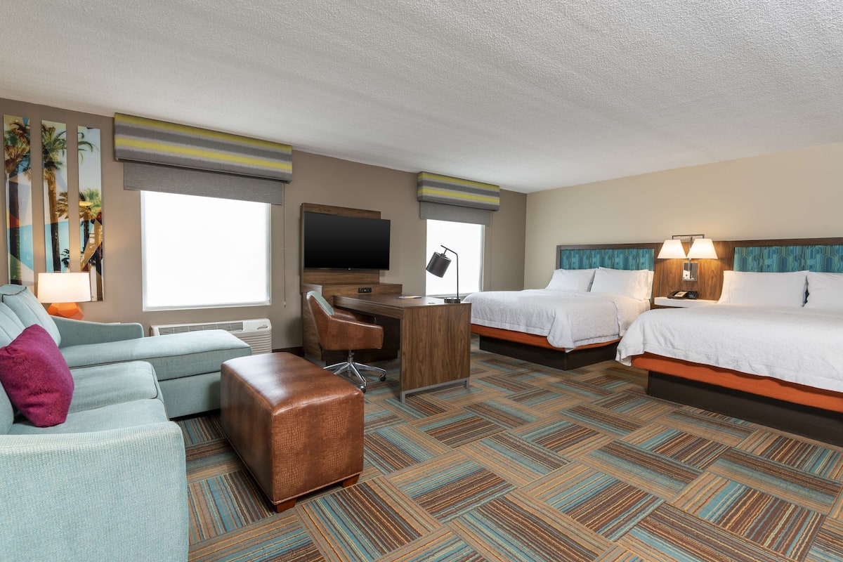 A hotel room with a sofa, two beds, a desk, a television, and a window with blinds.