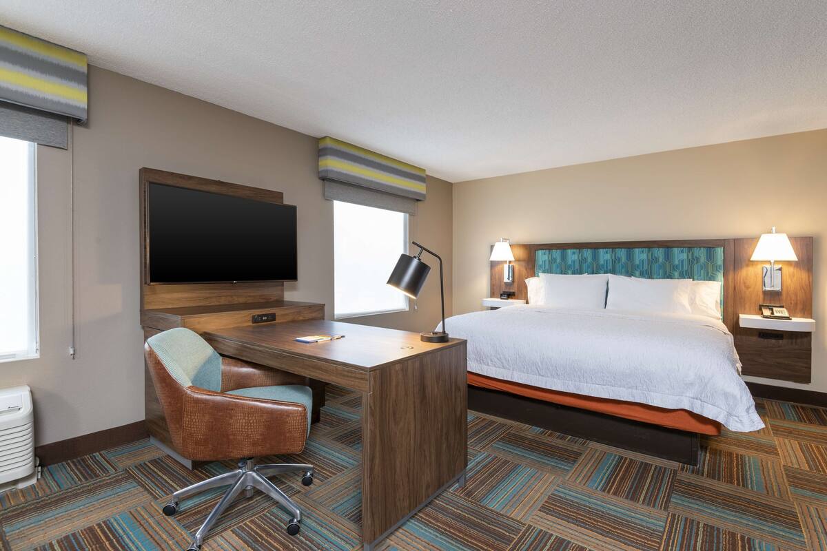 A hotel room with a bed, desk, chair, TV, lamps, and a patterned carpet.