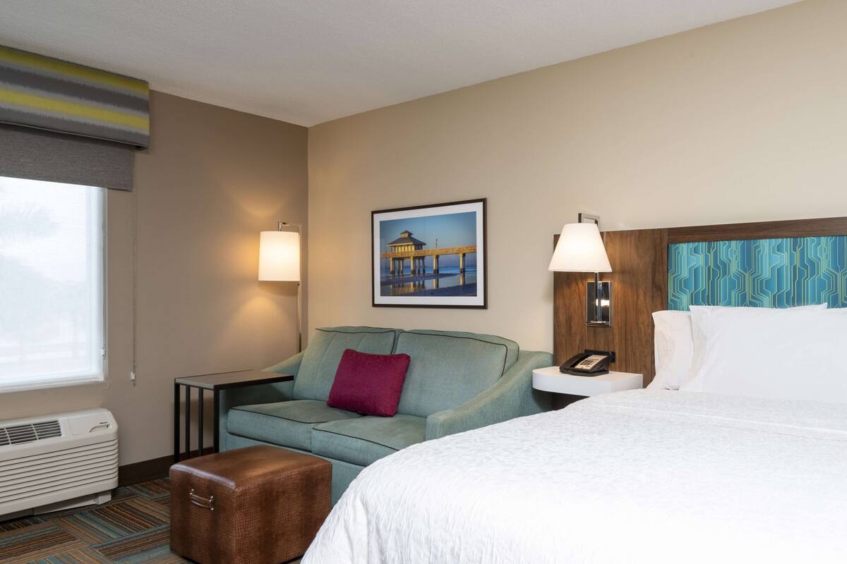 A hotel room with a bed, a sofa, a small table, a lamp, a picture on the wall, and a window with blinds.