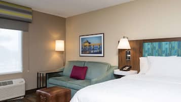 Quarto de hotel com cama, sofá, mesinha, luminária, quadro na parede e janela com persianas.