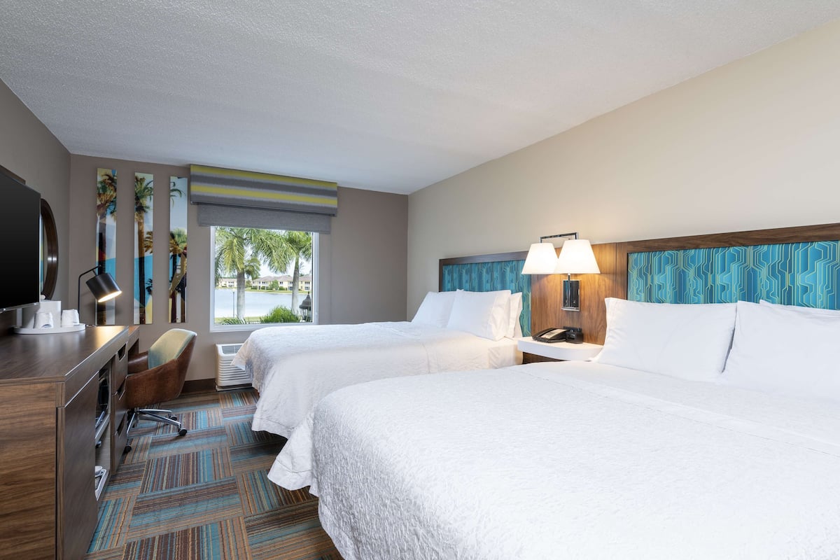 A hotel room with two beds, a desk, a chair, a TV, and a window with a view of palm trees.