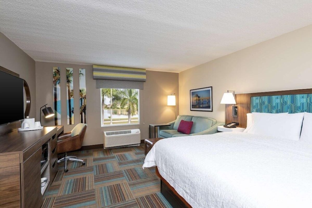 A hotel room with a large bed, a desk with a flat-screen TV, a chair, a lamp, and a window with a view of palm trees.