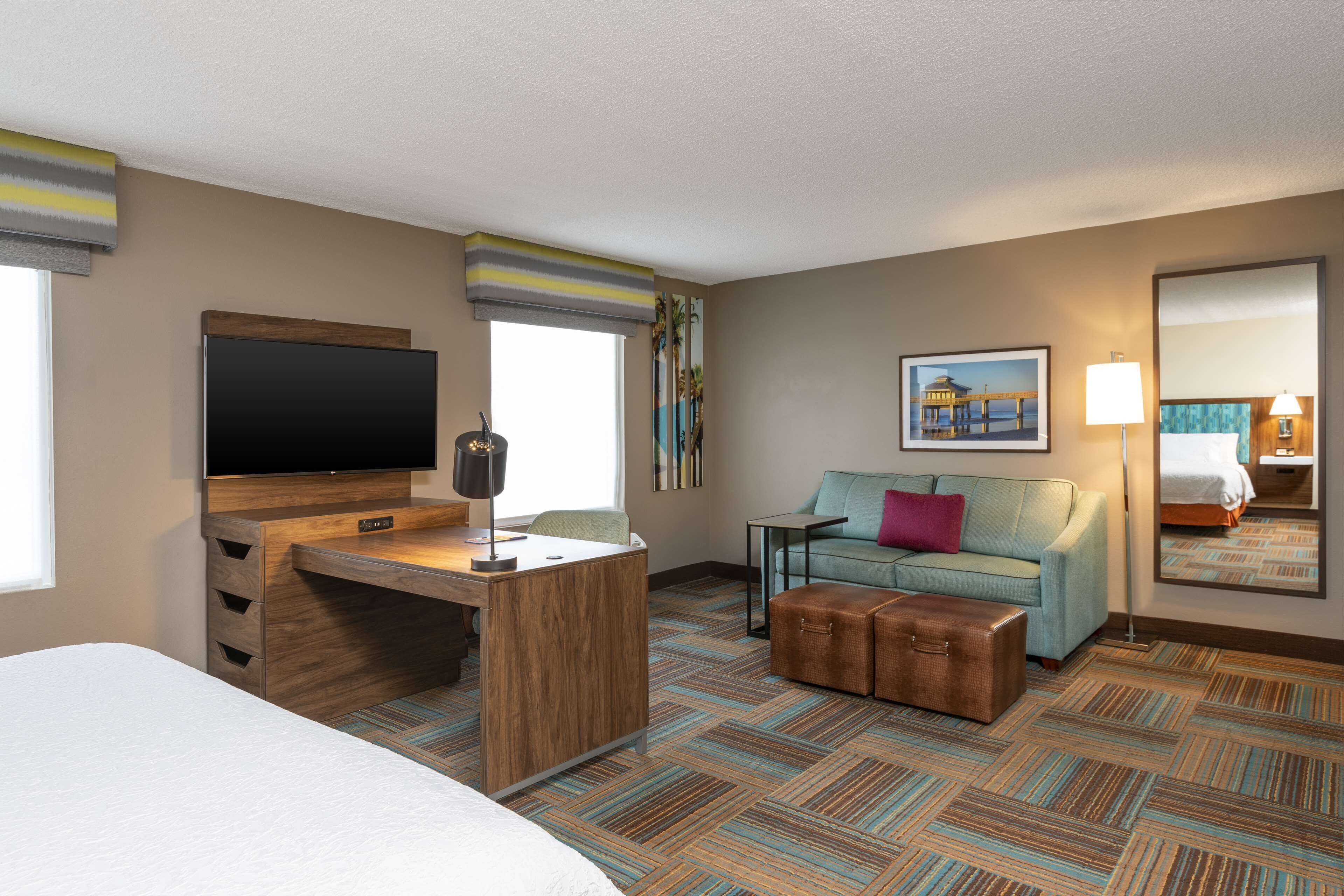 A hotel room with a bed, a desk with a flat-screen TV, a sofa, a small table, a suitcase, and a painting on the wall.