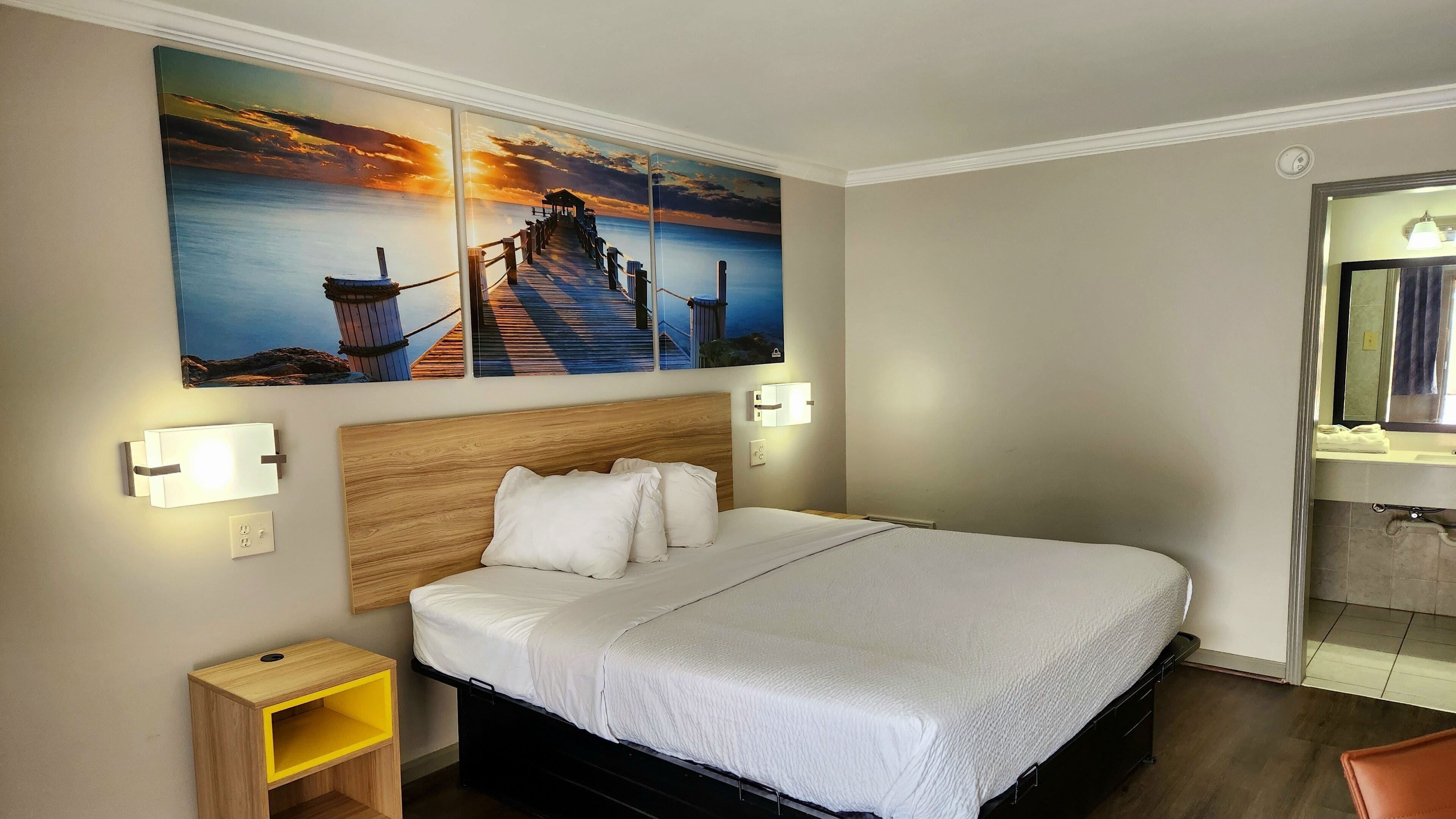 Camera d'albergo con un letto grande, due comodini, un tavolino e una testiera in legno, con un quadro raffigurante un tramonto su un molo.