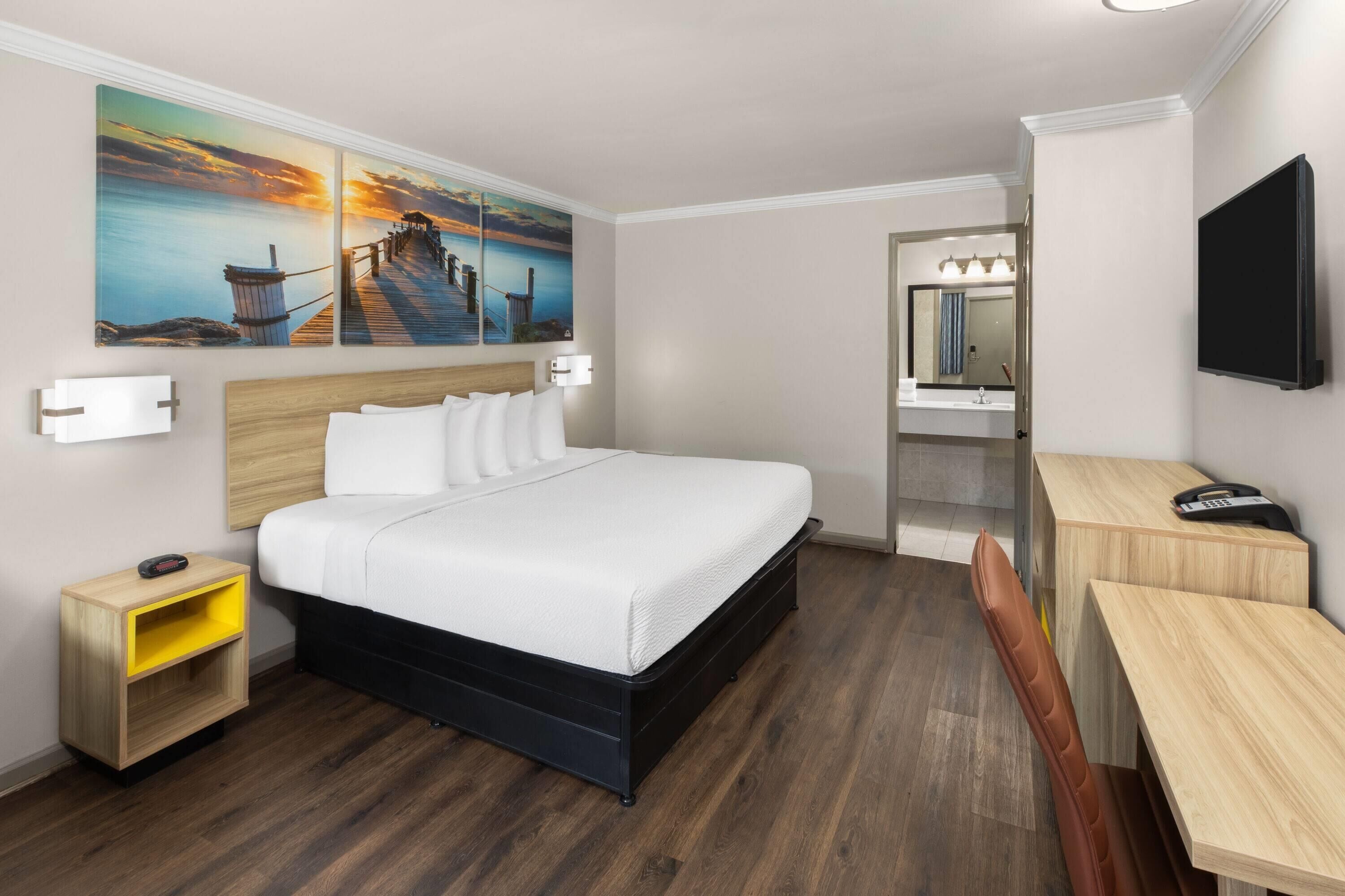 A hotel room with a bed, bedside table, desk, chair, and a painting of a sunset over water.