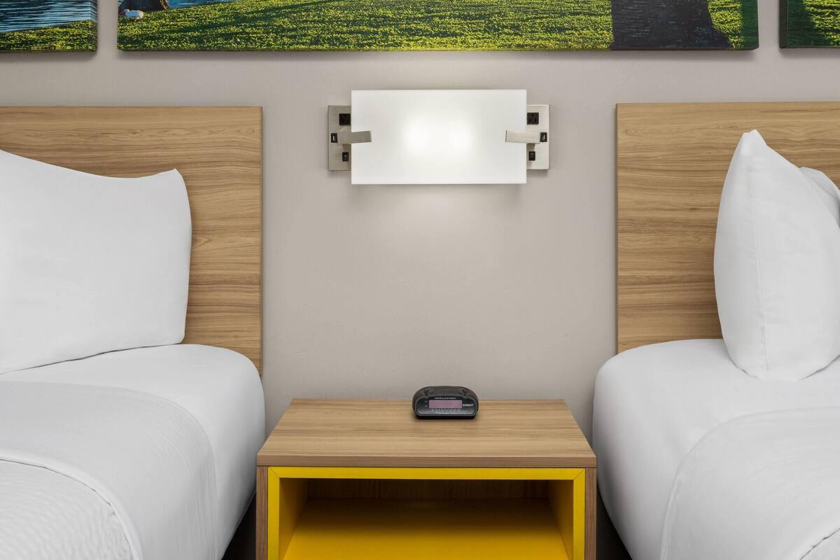 A hotel room with two beds, a bedside table with a clock, and a wall-mounted light fixture.