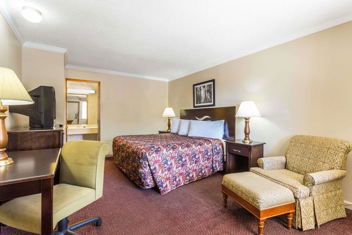 A hotel room with a bed, two chairs, a desk, a television, and a picture on the wall.