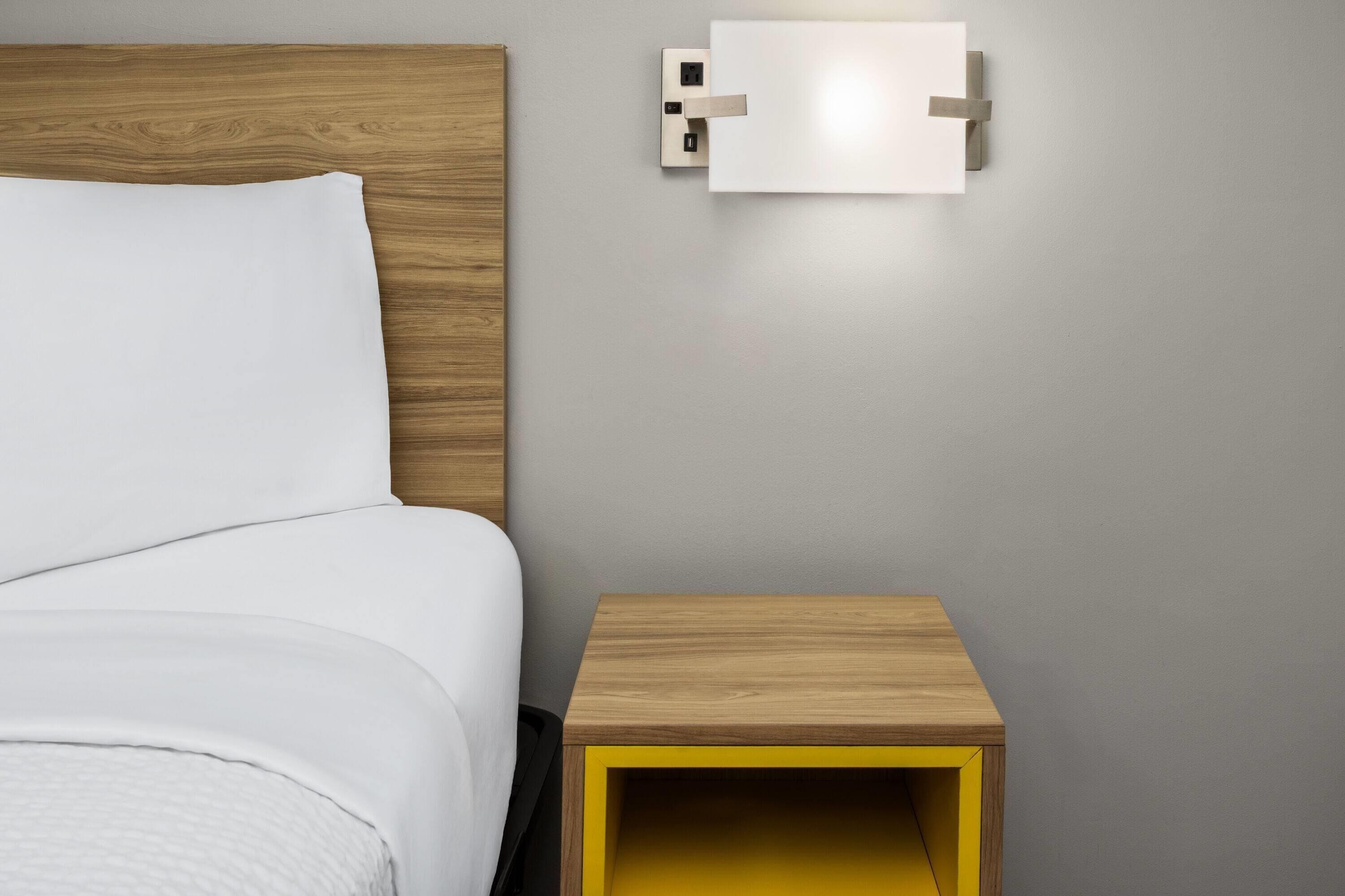 Un lit bien fait, avec du linge de lit blanc, une tête de lit en bois et une petite table de chevet en bois dotée d’un tiroir jaune.