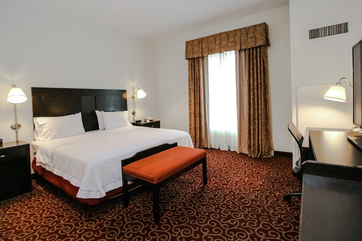 A hotel room with a large bed, two bedside tables, a desk with a chair, and a window with curtains.