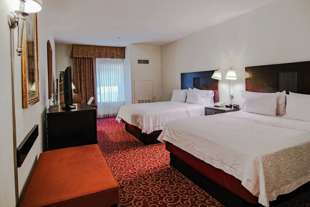 A hotel room with two beds, a desk, a chair, a TV, and a window with curtains.