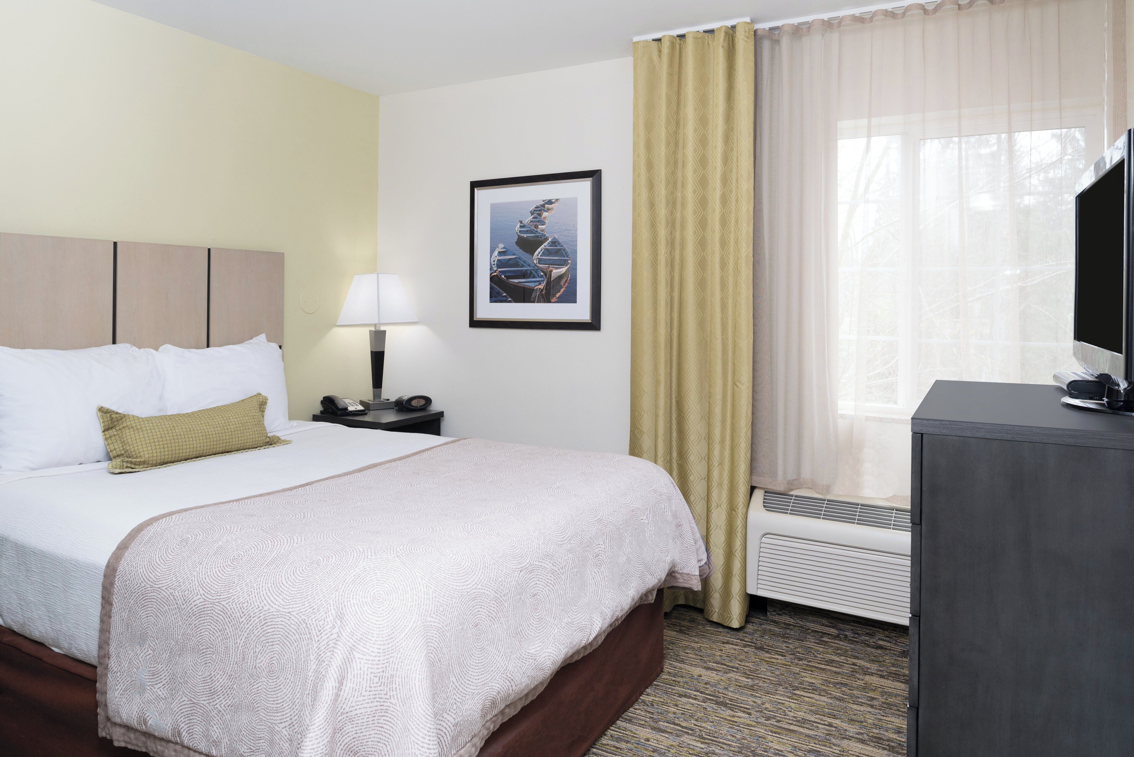 A hotel room with a bed, a bedside table with a lamp, a framed picture on the wall, and a window with curtains.