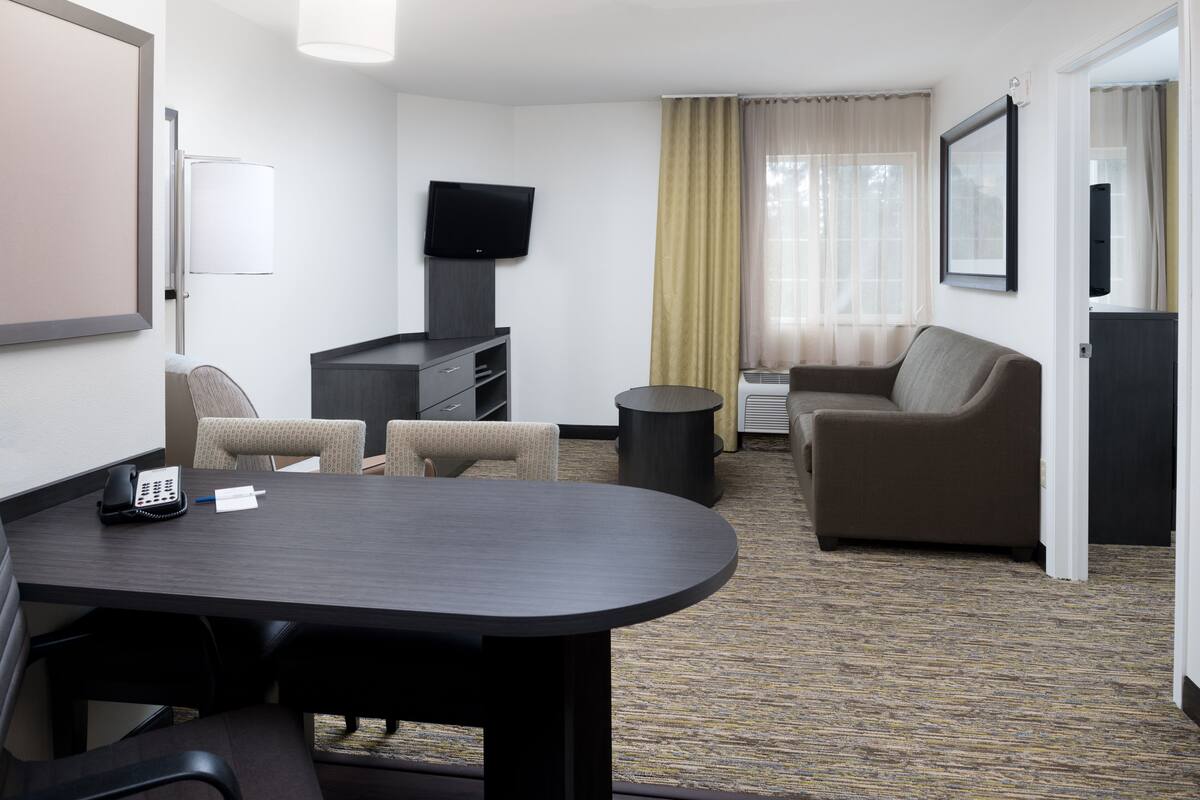 Ein Hotelzimmer mit Konferenztisch, zwei Sesseln, einer Couch, einem Fernseher, einem kleinen Mülleimer und einer Tür zu einem weiteren Raum.