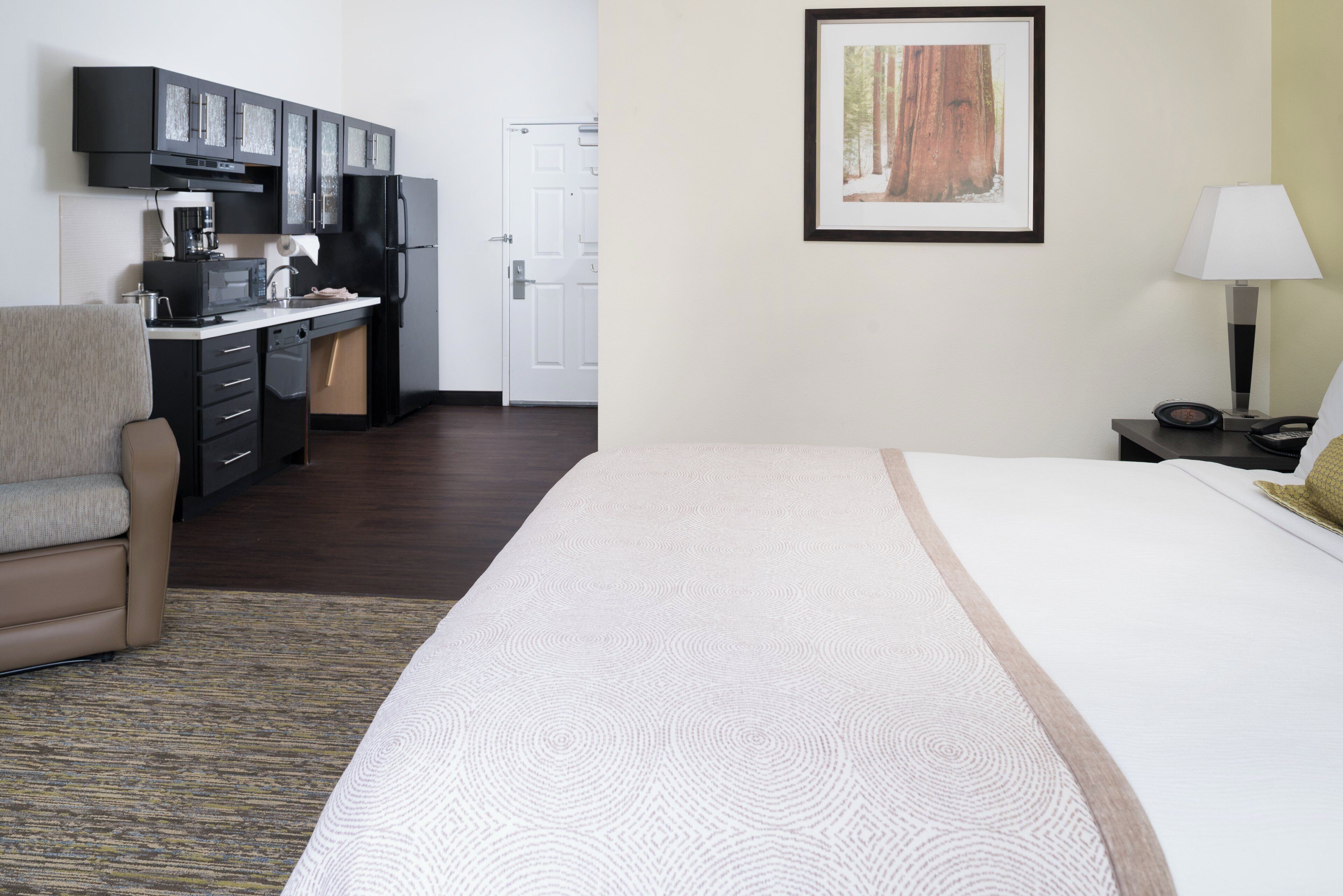 A hotel room with a bed, a chair, a desk with a computer, a kitchen area, and a framed picture on the wall.