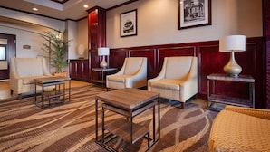 Un hall d’hôtel doté d’un sol recouvert de moquette, de trois fauteuils, d’une table basse, d’une table d’appoint avec une lampe, d’une plante en pot et de tableaux encadrés accrochés au mur.