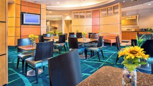 Un hall d’hôtel doté de plusieurs espaces de détente, comprenant des tables et des chaises, un téléviseur fixé au mur et un coin cuisine à l’arrière-plan.