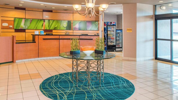 Un hall d’hôtel comprenant un comptoir de réception, une table avec un plateau en verre et un vase, des plantes en pot, un distributeur automatique et une grande fenêtre.