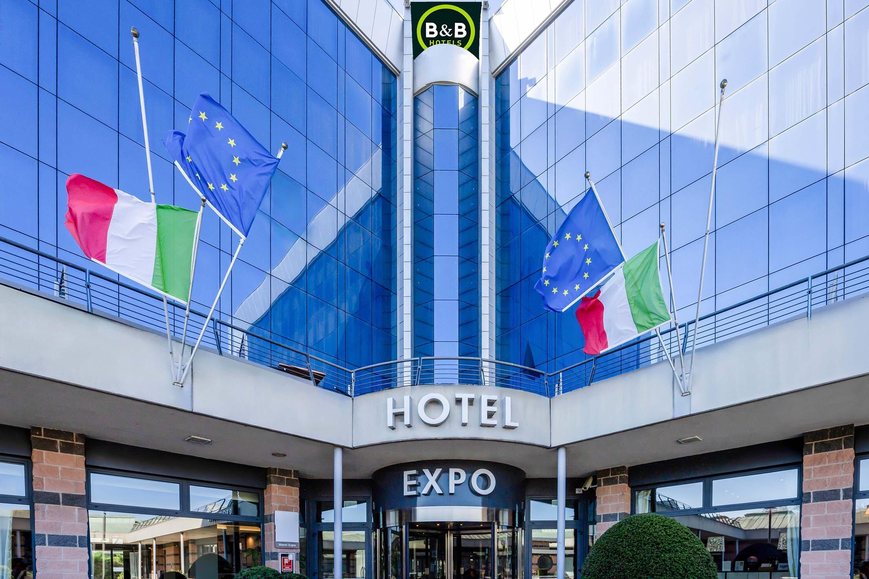Een modern hotel met een glazen gevel, vlaggen van de EU, Italië en het Verenigd Koninkrijk, en een bord waarop 'HOTEL EXPO' staat.