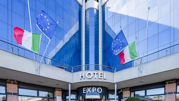 Un hotel moderno con fachada de vidrio, banderas de la Unión Europea, Italia y el Reino Unido, y un letrero que dice 'HOTEL EXPO'.