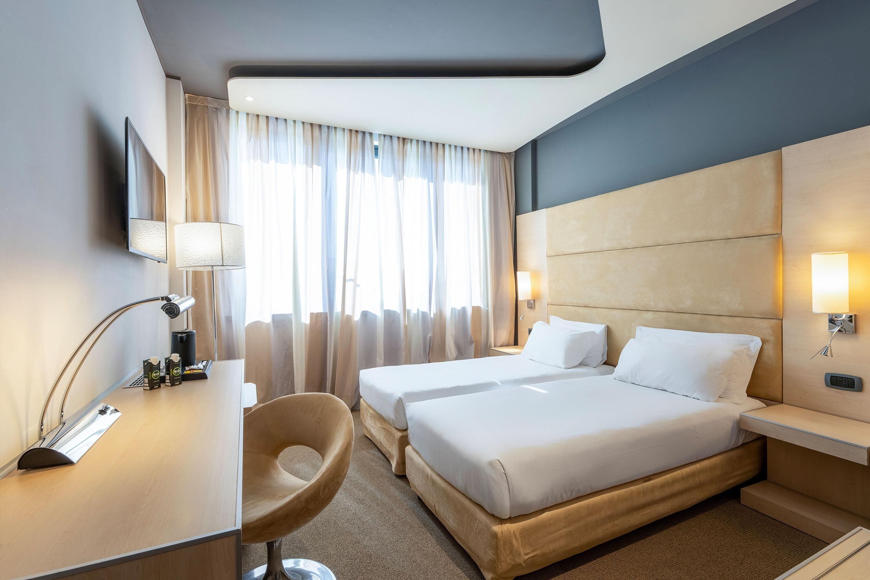 Habitació d'hotel amb dos llits, un escriptori, una cadira, una làmpada i una finestra amb cortines.