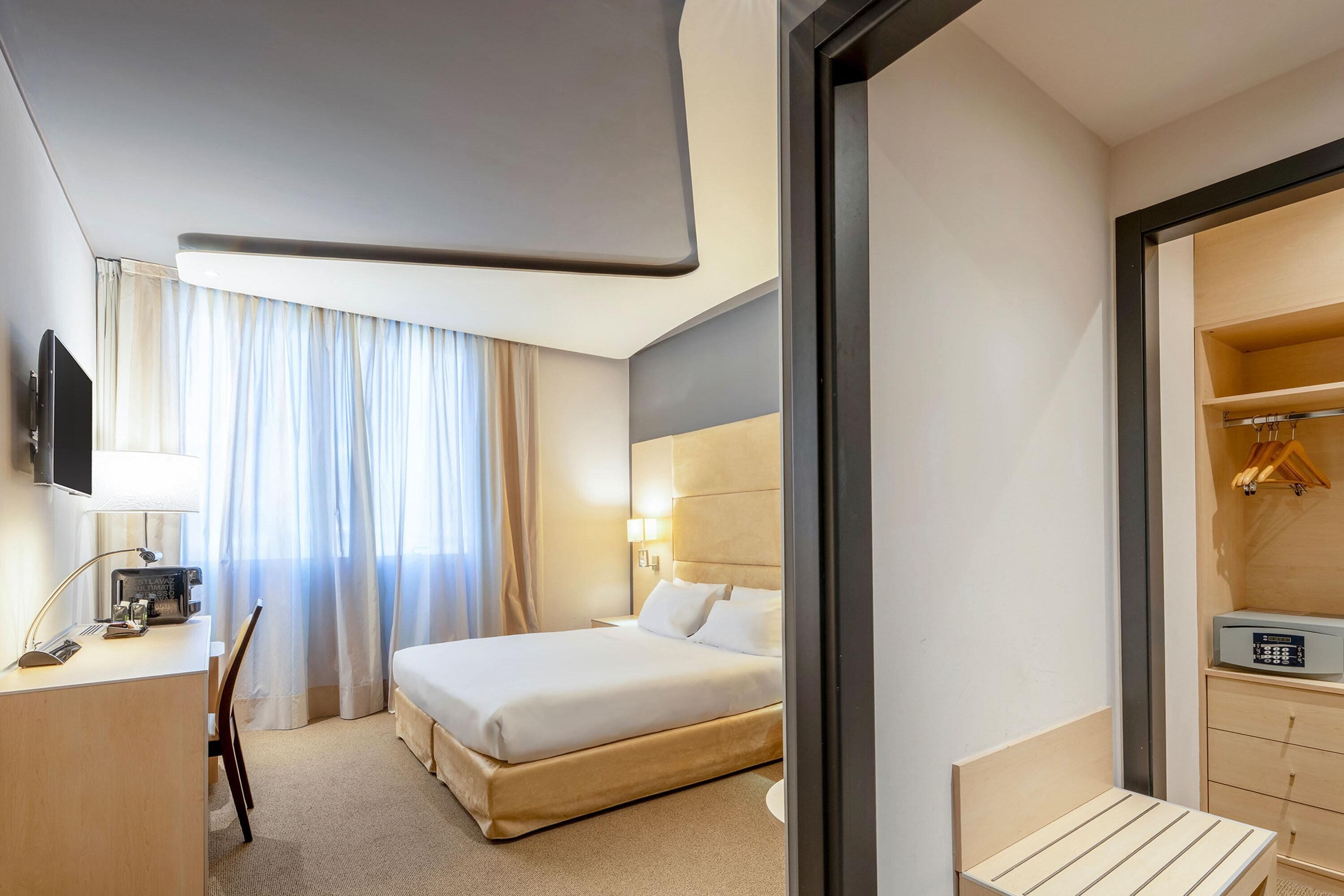 Habitación de hotel con cama, escritorio con lámpara, televisor y armario.