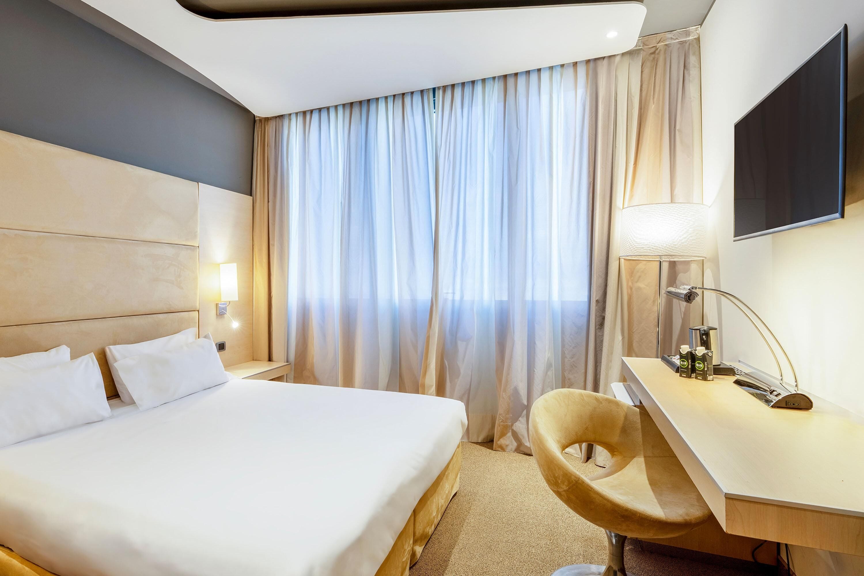 Ein Hotelzimmer mit Bett, Schreibtisch, Stuhl, Fernseher und einem Fenster mit Vorhängen.