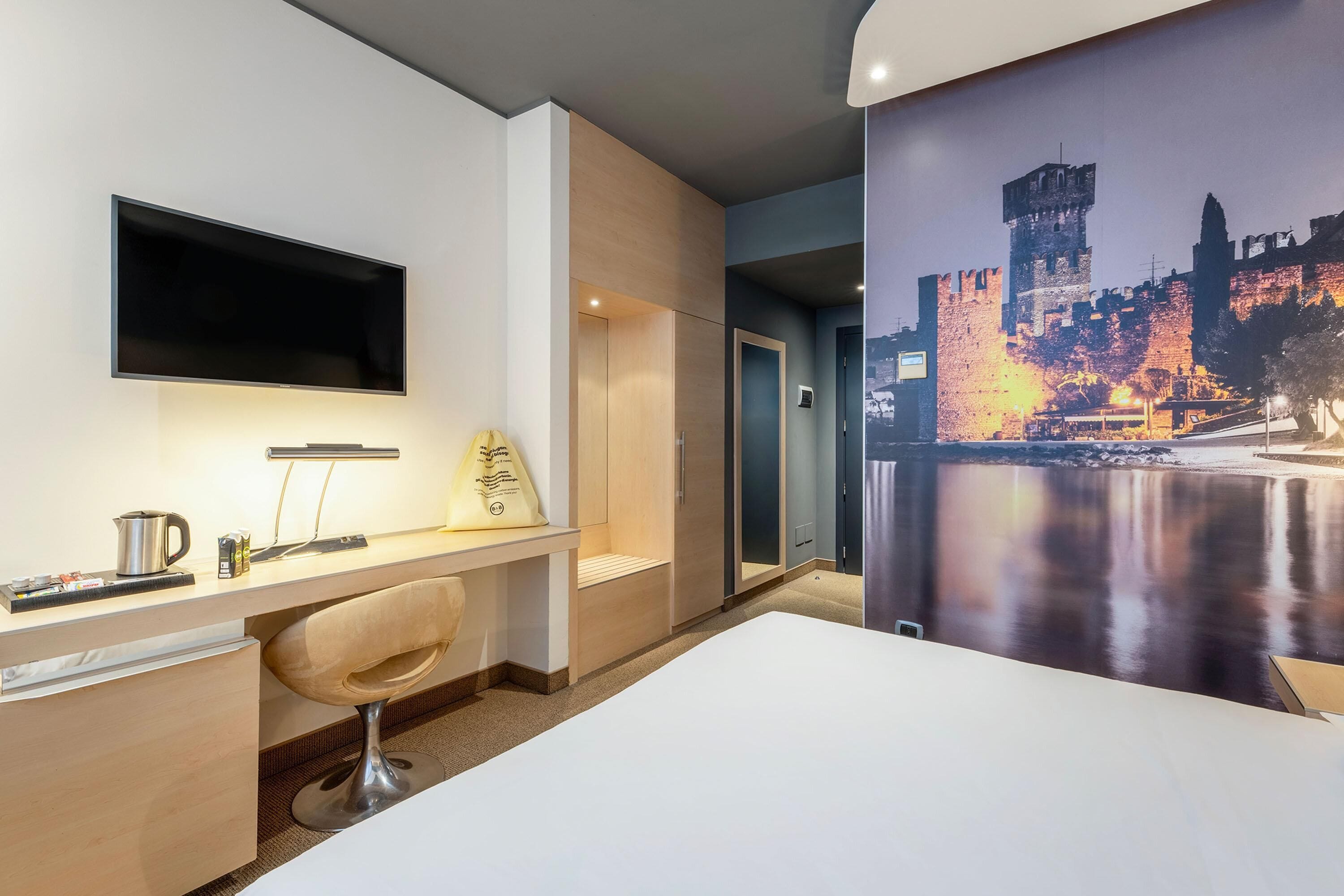 A hotel room with a bed, a desk with a chair, a flat-screen TV, and a painting of a castle.