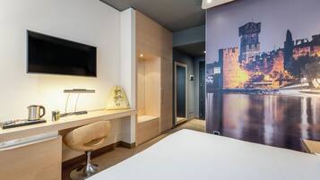 A hotel room with a bed, a desk with a chair, a flat-screen TV, and a painting of a castle.